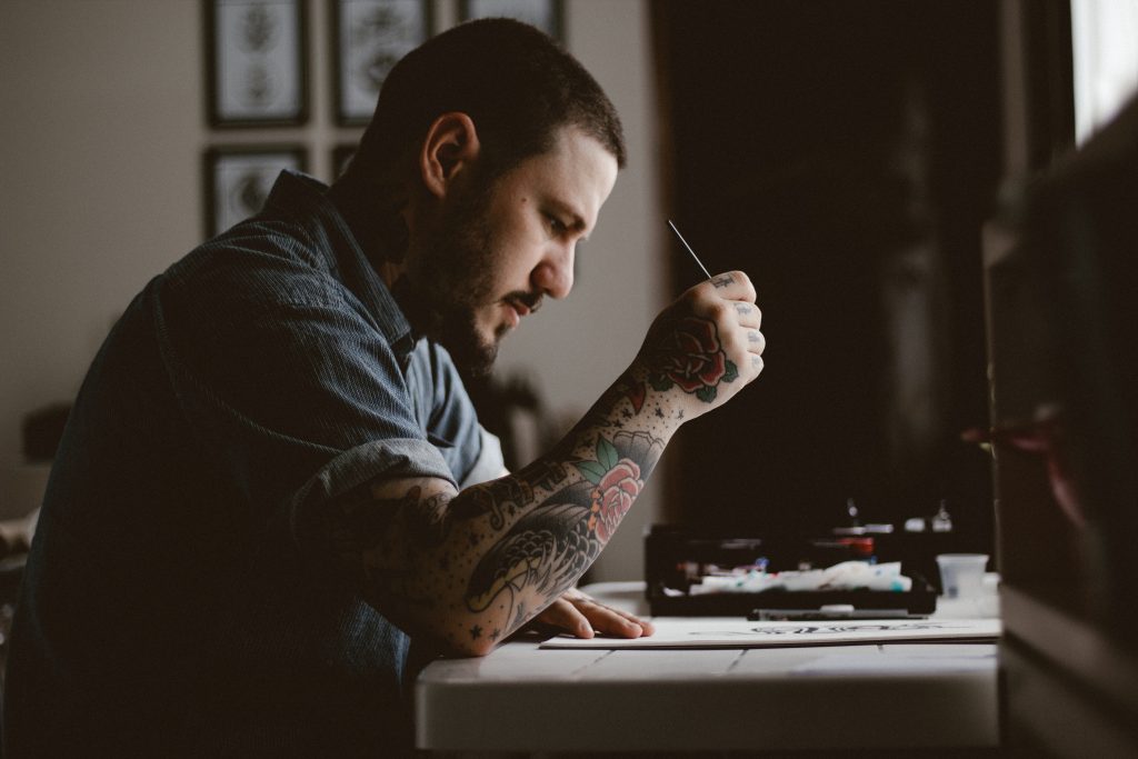 Man with tattoos on his arm drawing at a desk.