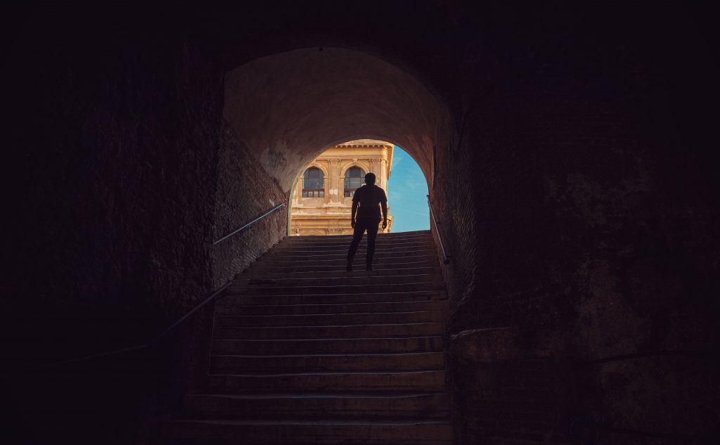 Silhouette of a man standing near the top of a stone staircase, with a blue sky at the top. 