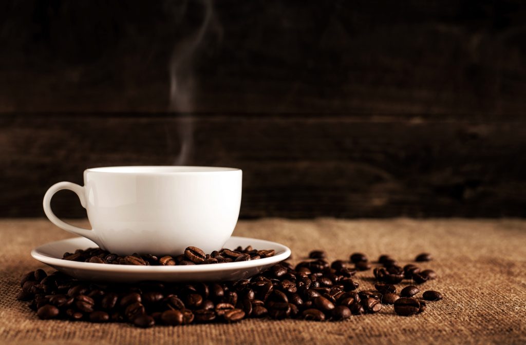 White coffee cup with steam coming out of it and cup is surrounded by coffee beans.