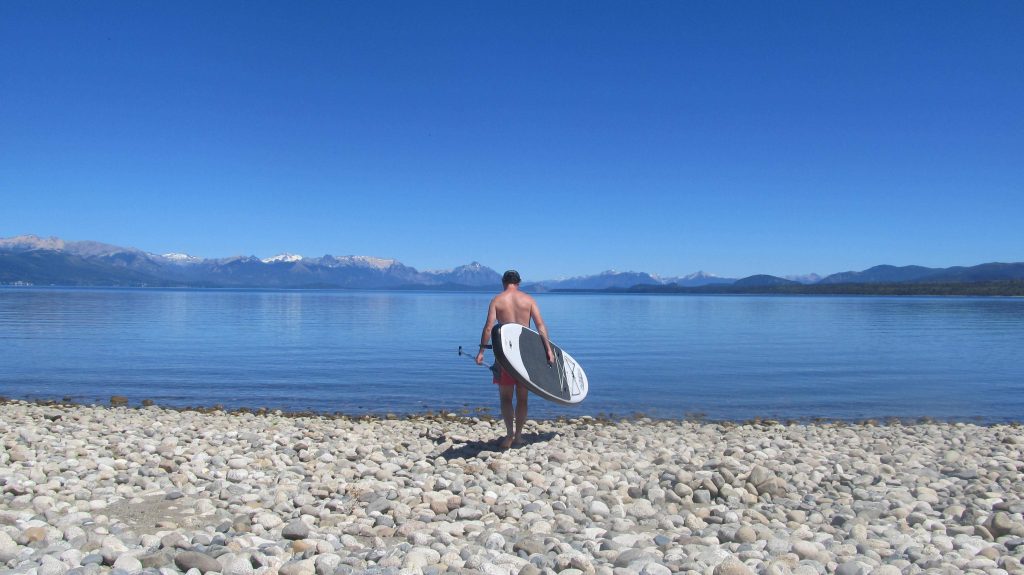 A man with a paddle board on the shore of a lake, about to walk in.