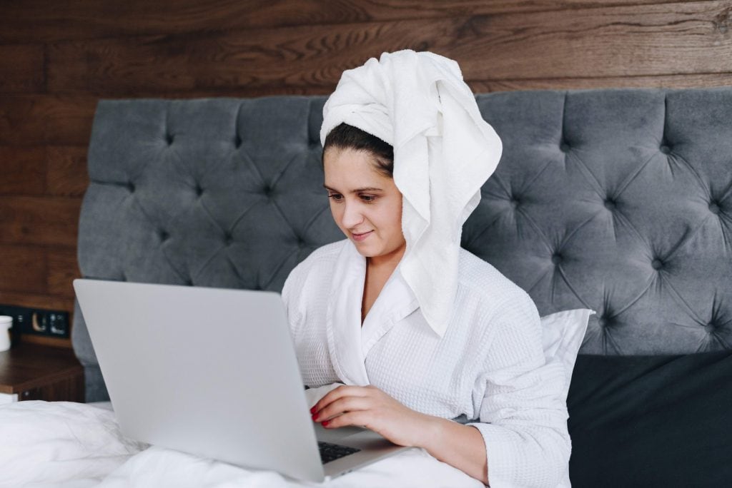 Woman in dressing gown and towel around her head working on a laptop while sitting on a bed.