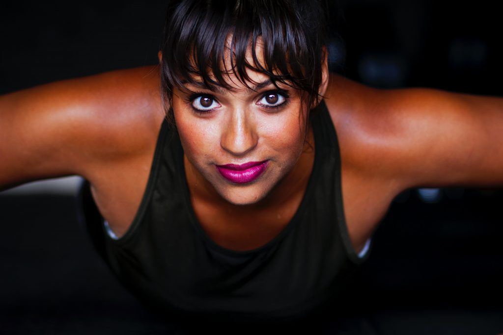 Dark-skinned young woman in black vest doing push-ups while smiling at the camera.