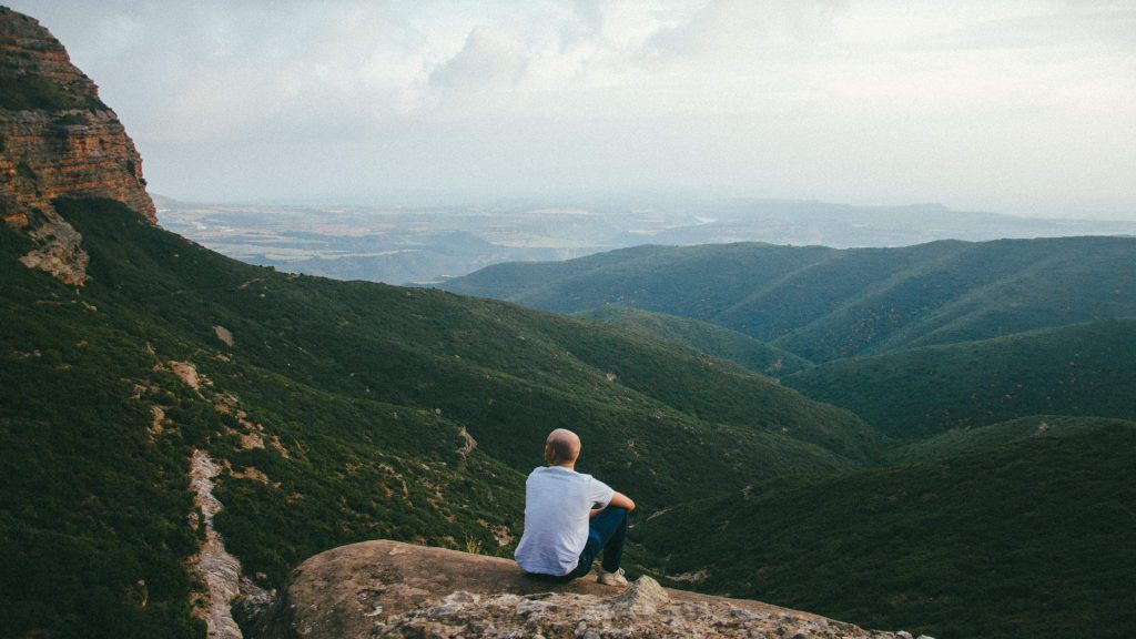 Man sitting on a cliff edge looking over a mountain range.