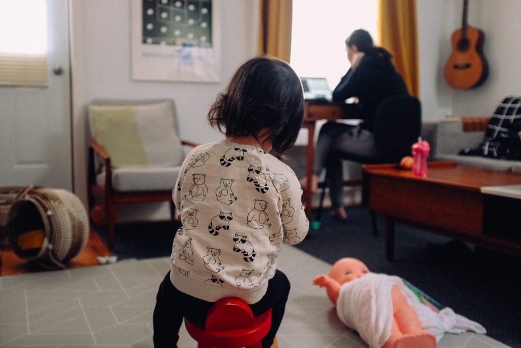 Women works on laptop at home with young child in the foreground.