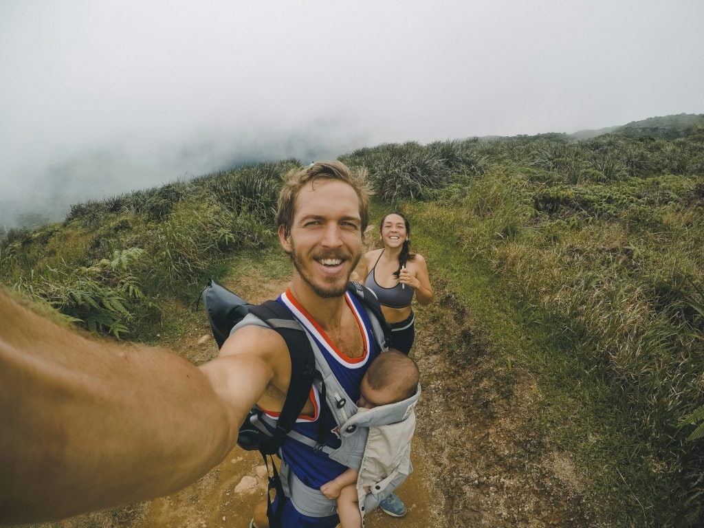 Selfie of a smiling man with a baby and a woman in the background, exercising on a grassy mountain.