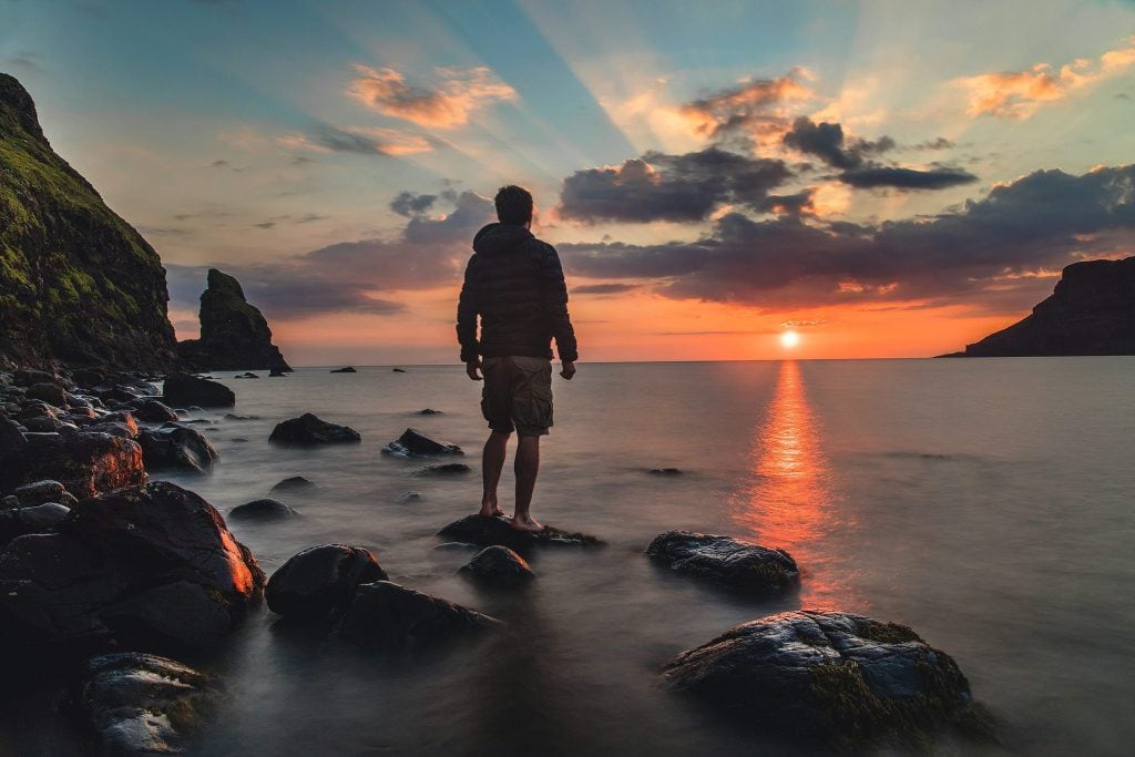 Man standing on a rock on the shore of the sea, looking at the sunrise.