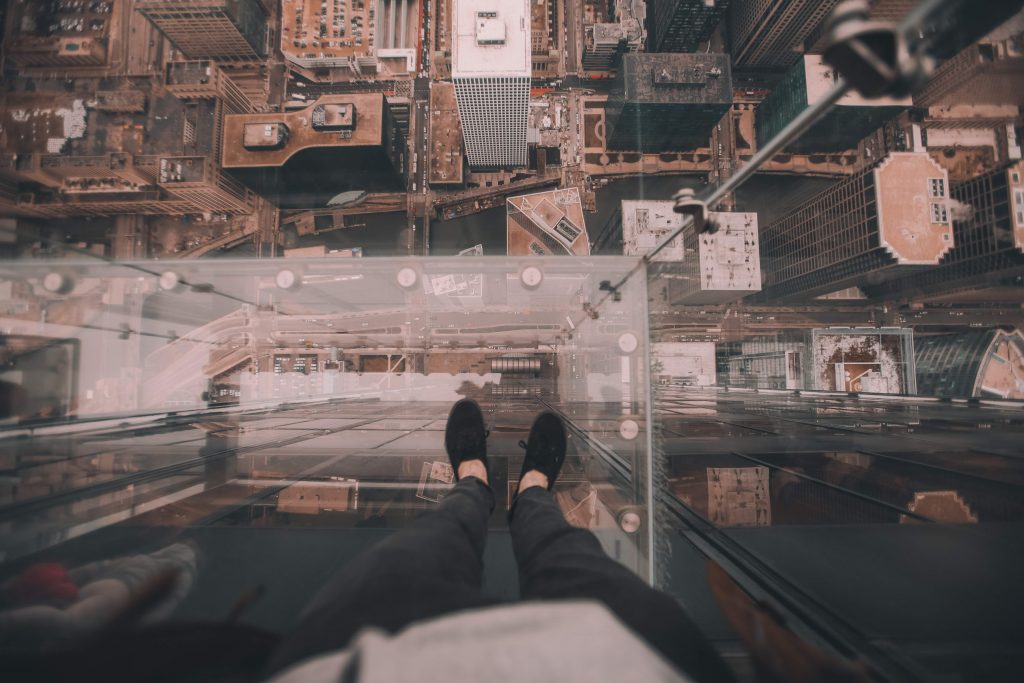 First person view of man's legs and feet standing on glass ledge of highrise, looking down at the streets and buildings far below.