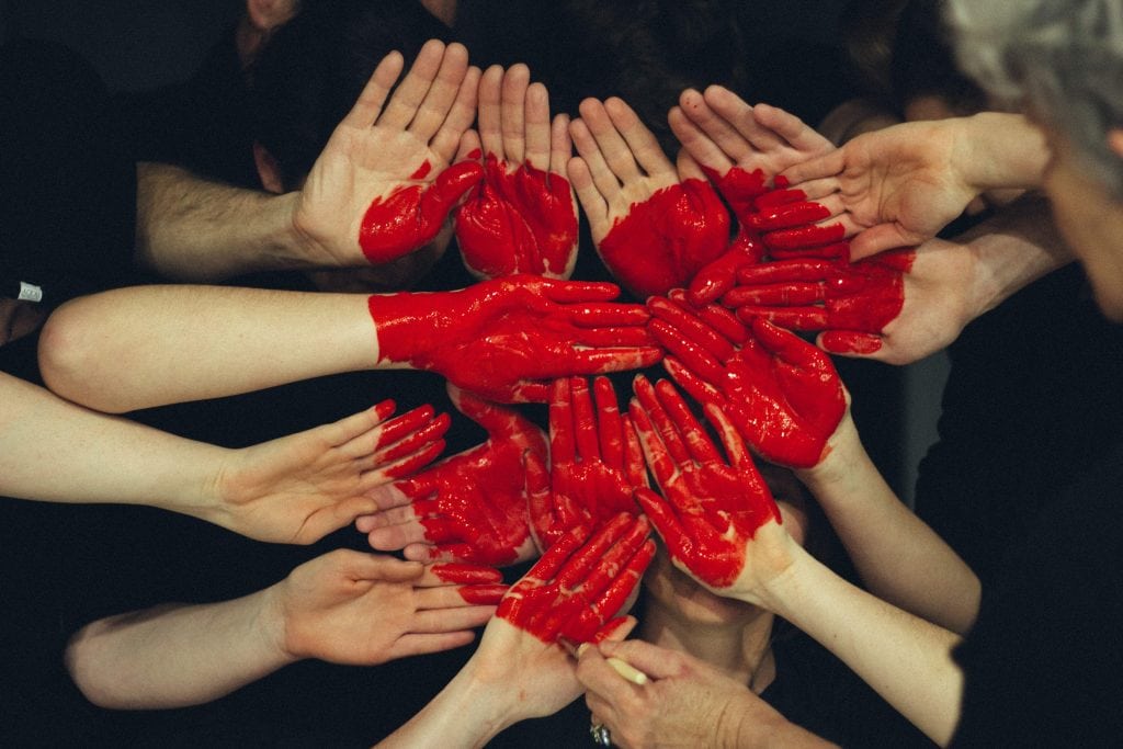 A group of hands gathered together with a red heart painted over them.