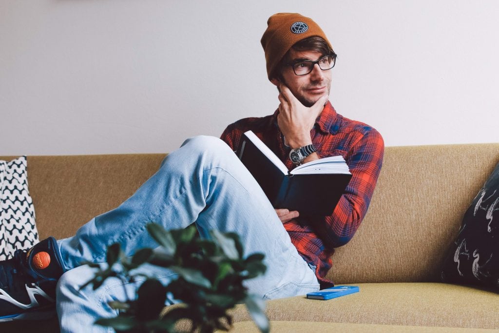 Man in glasses and beanie sitting on a sofa reading a book.