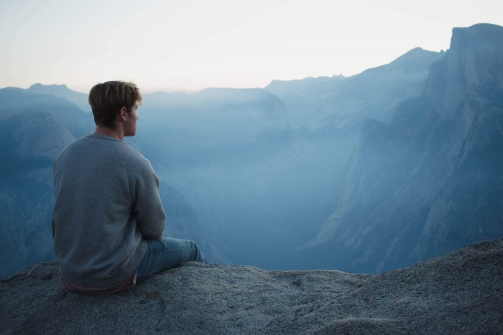 Man sitting on cliff edge, looking at mountain range at dusk.