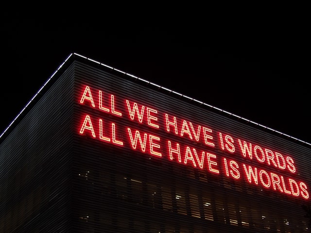 Large red neon sign on a building saying 'All we have is words; All we have is worlds'.