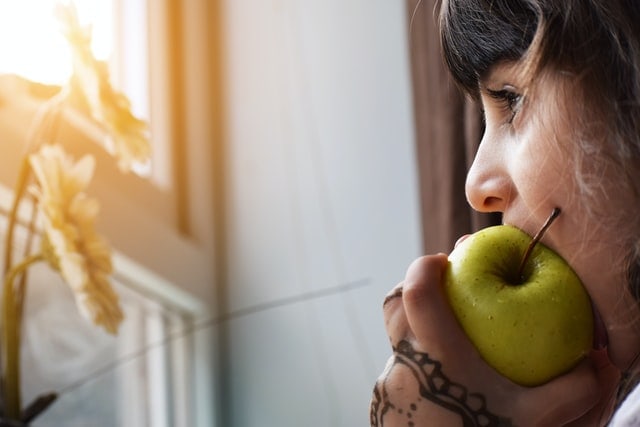Young girl eating a green apple while looking out the window.