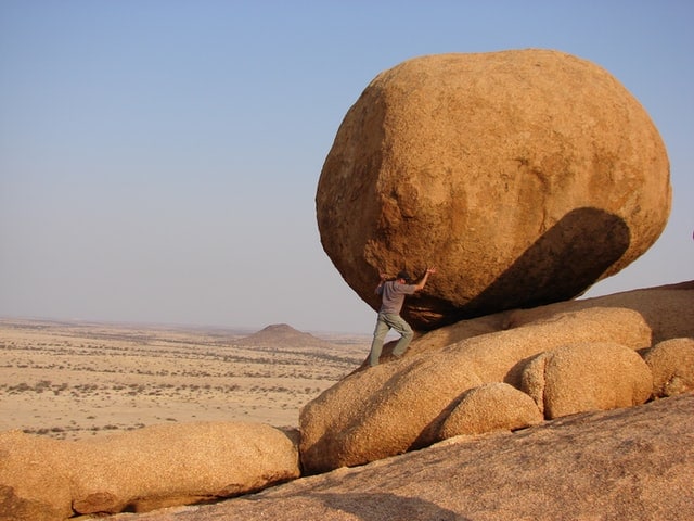 Man seemingly holding up a large boulder in a desert.
