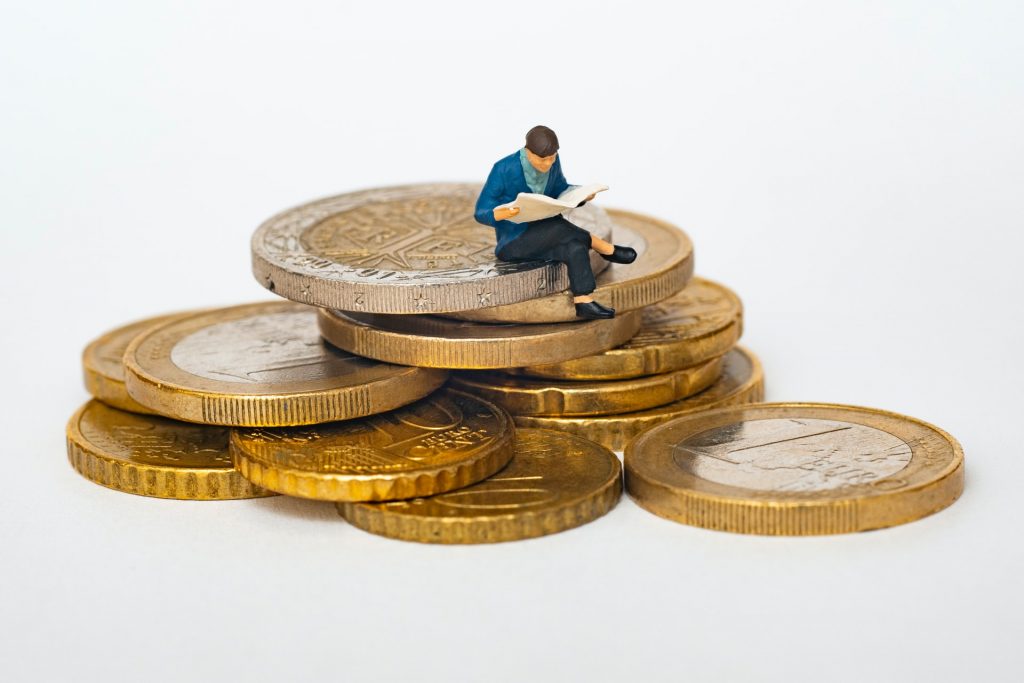 Tiny plastic model of a man reading a newspaper, sitting on top of a pile of actual coins.