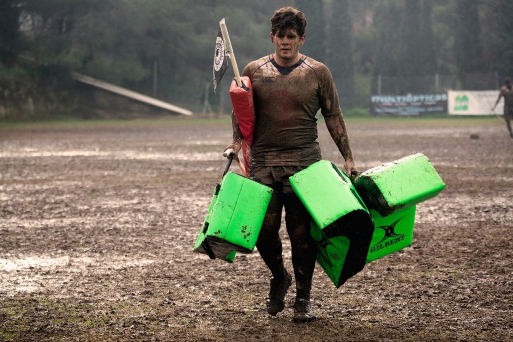 Man covered in mud carrying rugby practice equipment.