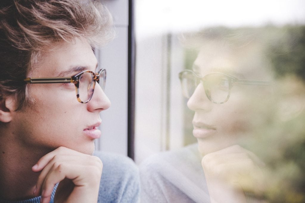 Young man in glasses is looking thoughtfully through a window while his reflection shows in the glass.
