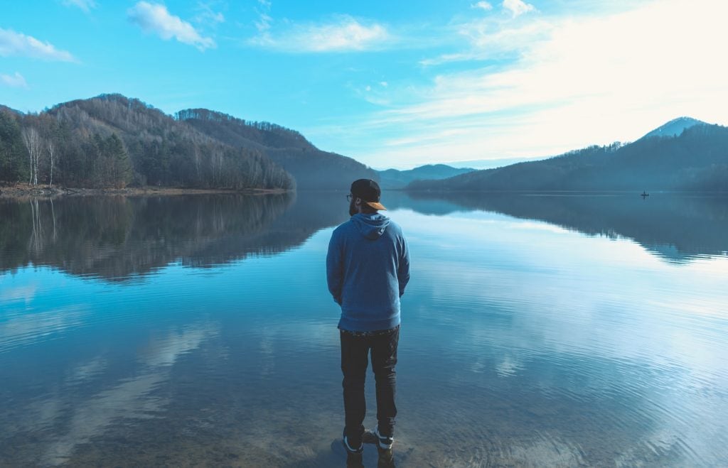 Man standing at the foot of a big lake with mountains in the distance.