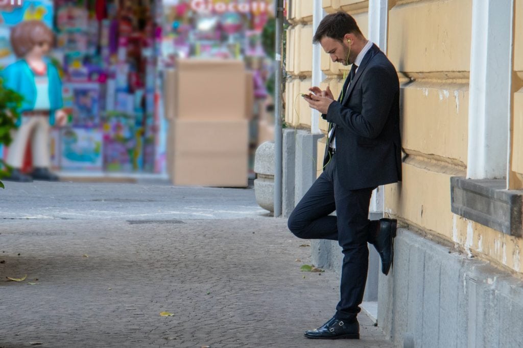 Man in a suit, leaning against a wall while looking at his smartphone.