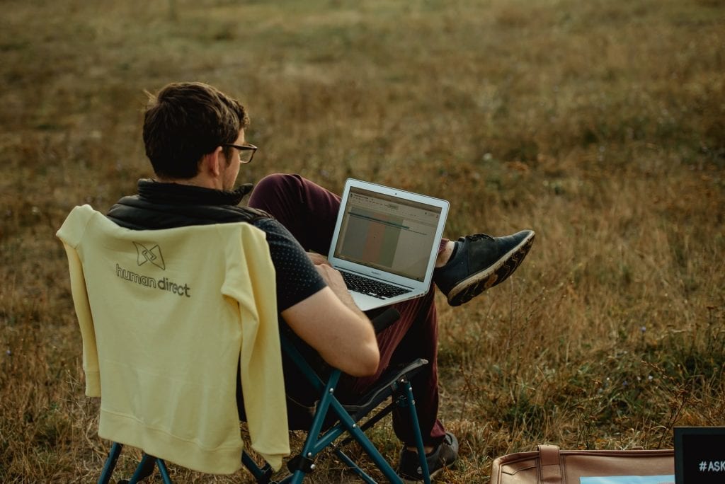 Man sitting in camping chair in field while working on laptop.
