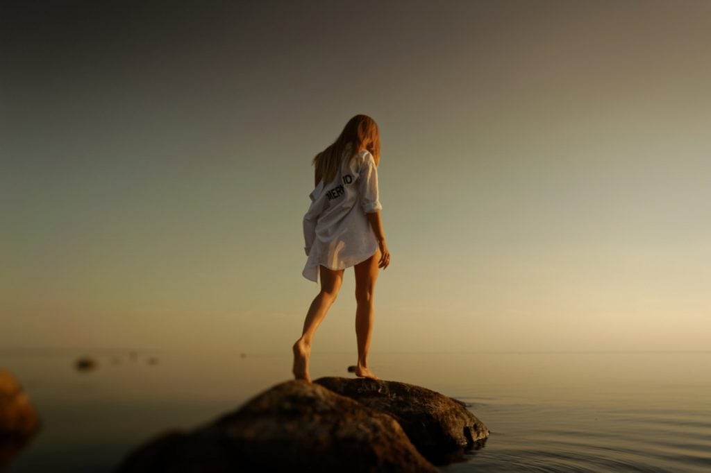 Back view of a woman in a dress standing on a rock overlooking a lake at sunset.