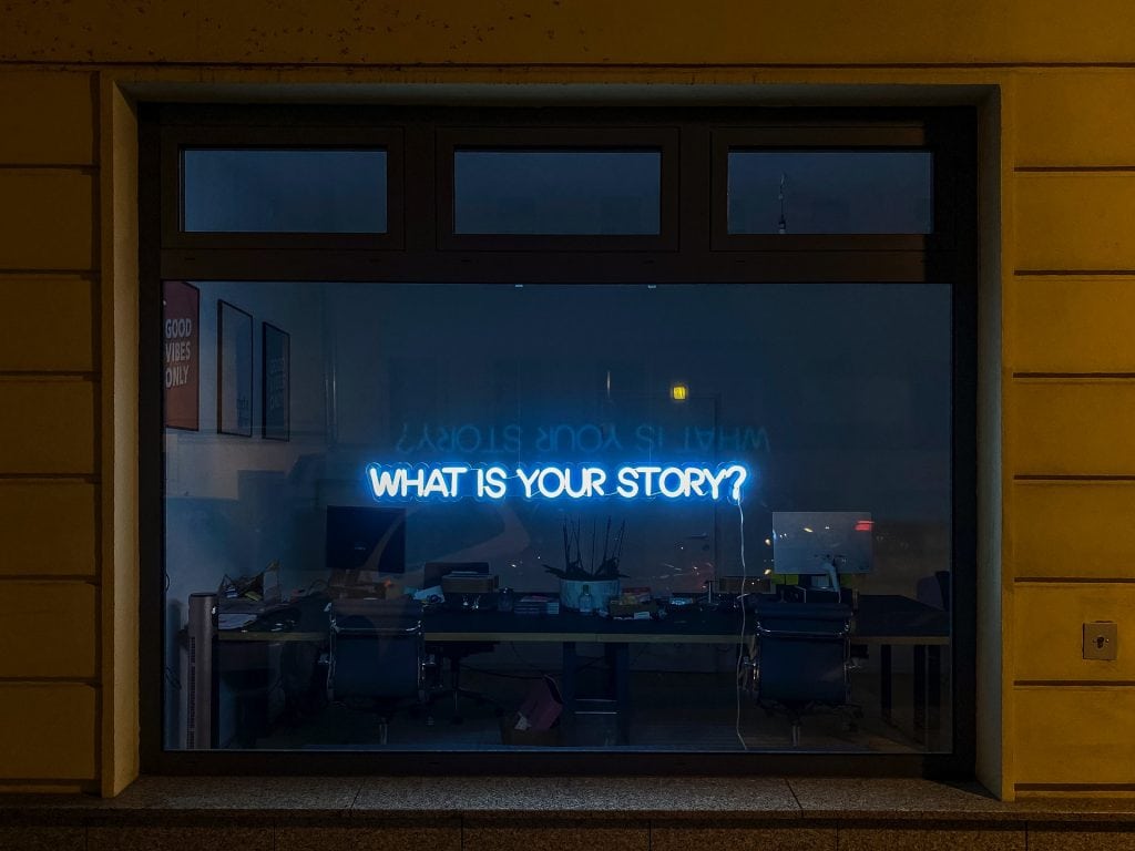 window with neon sign saying 'what is your story?'